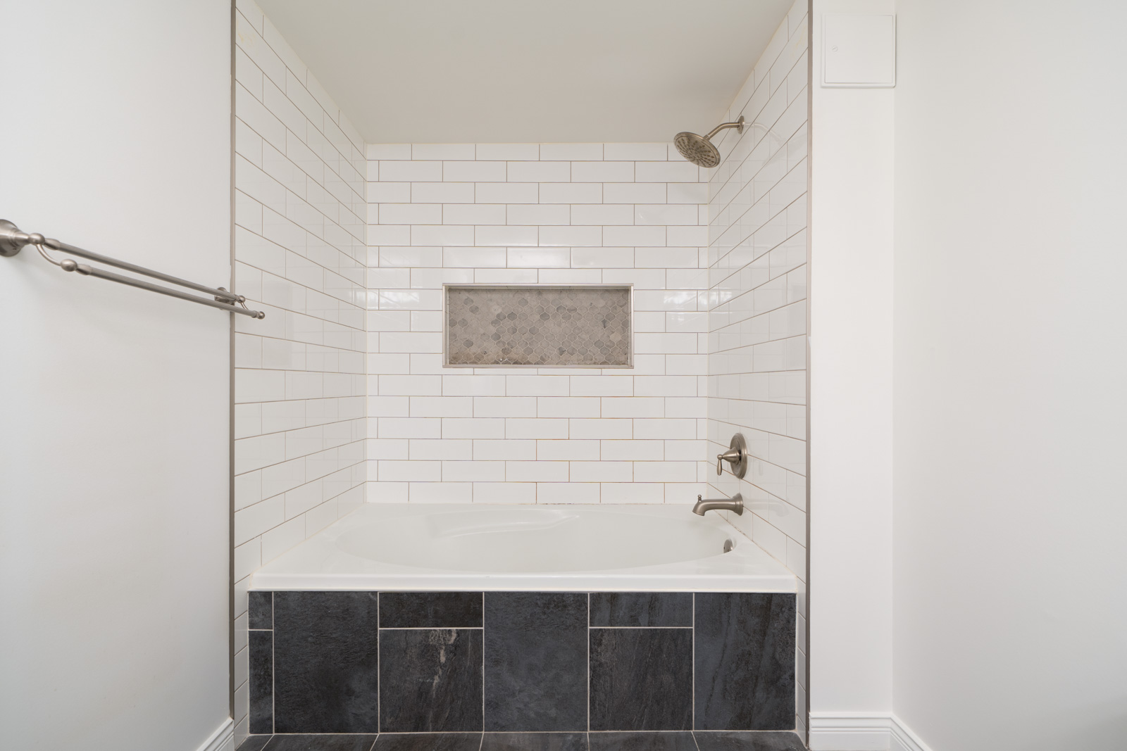 White-tiled tub and shower with built-in shelf, dark lower tiles, metal fixtures, and white walls at 1245 Quayside Dr, New Westminster.