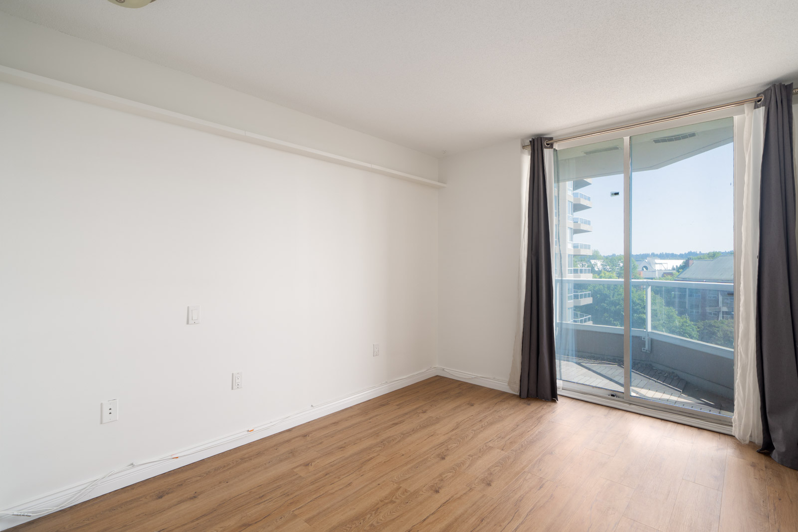 Empty room with white walls, wood floor, and large windowed door to a balcony; cityscape and trees seen from 12th floor, New Westminster.