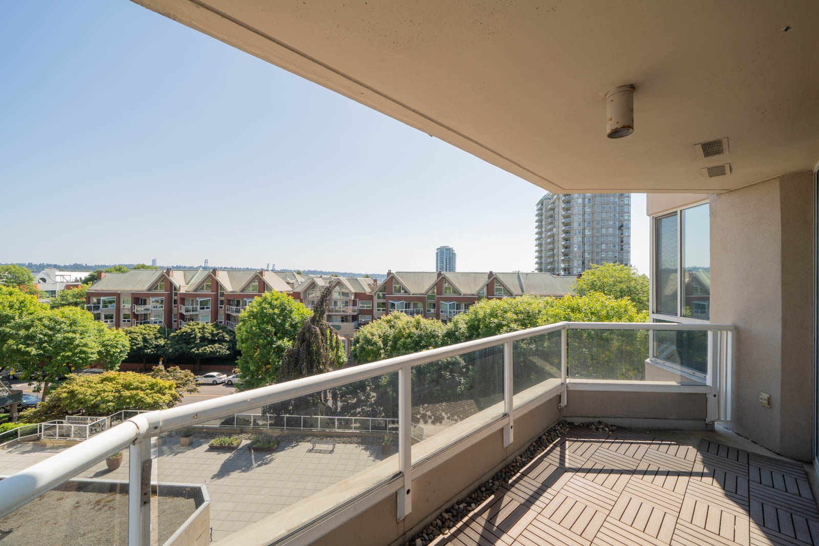 Balcony with glass railing overlooks a tree-lined street, townhouse roofs, and high-rises under clear sky in New Westminster.