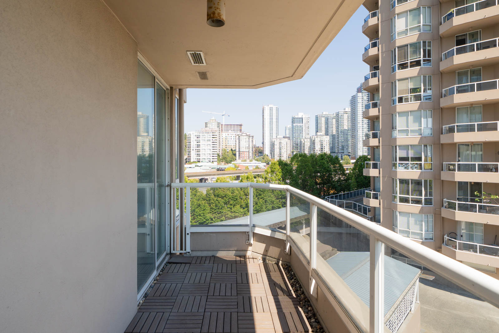 12th-floor apartment balcony in New Westminster with wood tile flooring, glass railing, and views of high-rise city buildings.