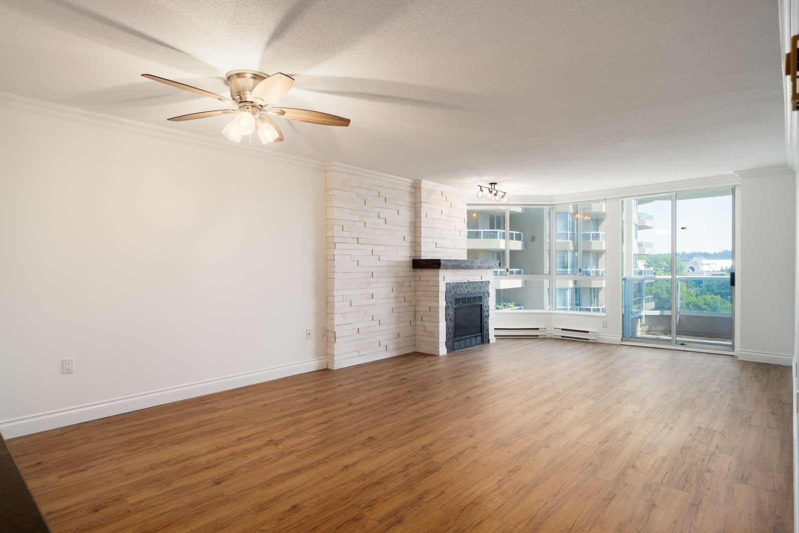 Empty living room with wood floors, white walls, ceiling fan, fireplace, and balcony windows on the 12th floor in New Westminster.