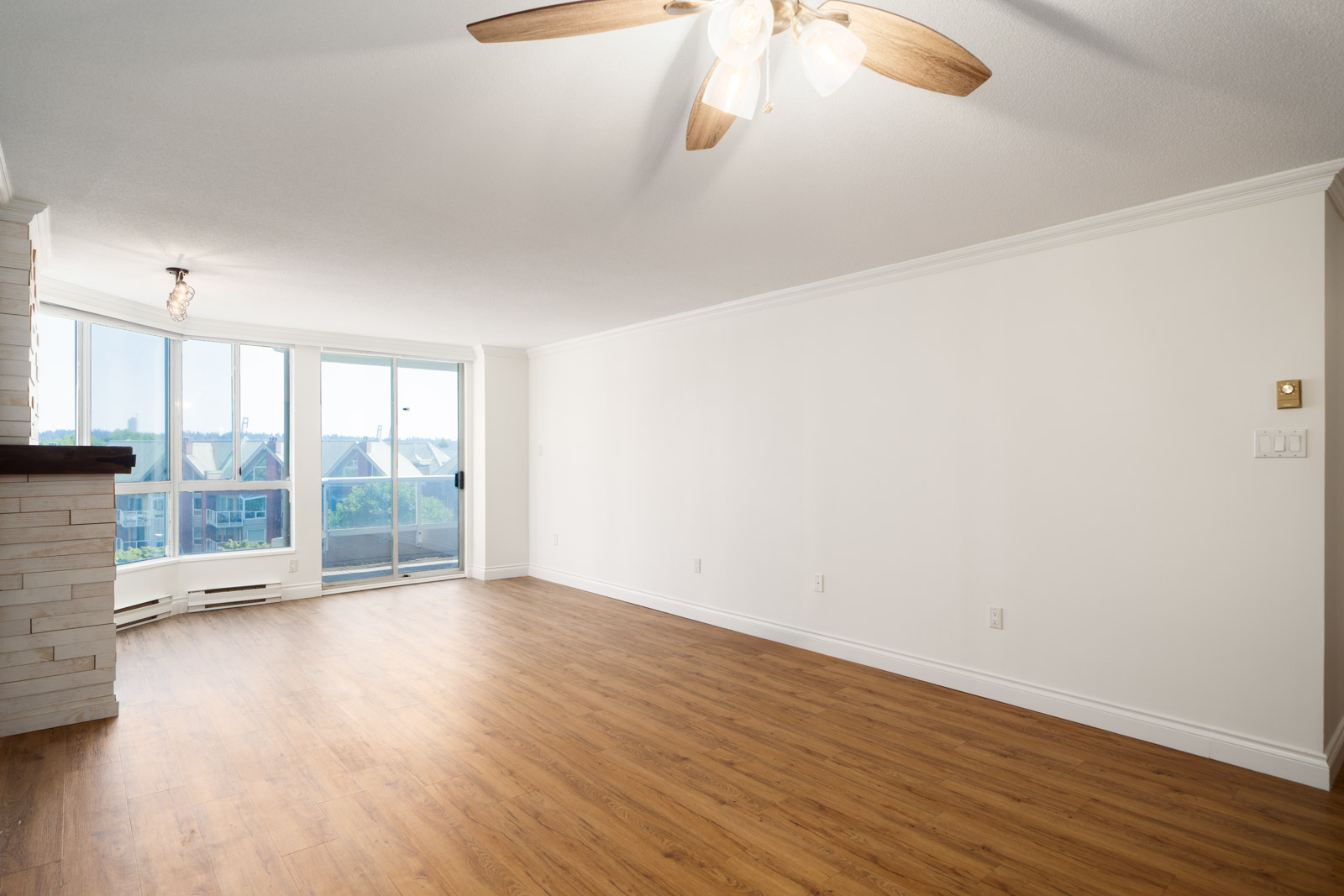 Bright, empty living room in New Westminster with light wood floors, white walls, ceiling fan, large windows, and balcony door.
