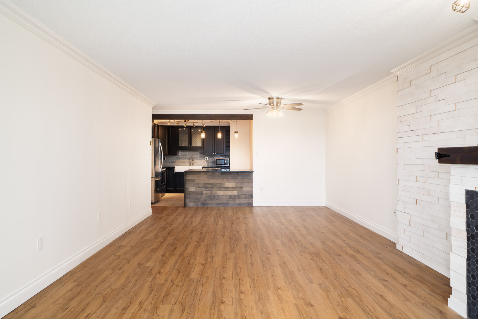 Spacious empty living room with wood floors, white walls, stone fireplace, open to modern kitchen in New Westminster condo.