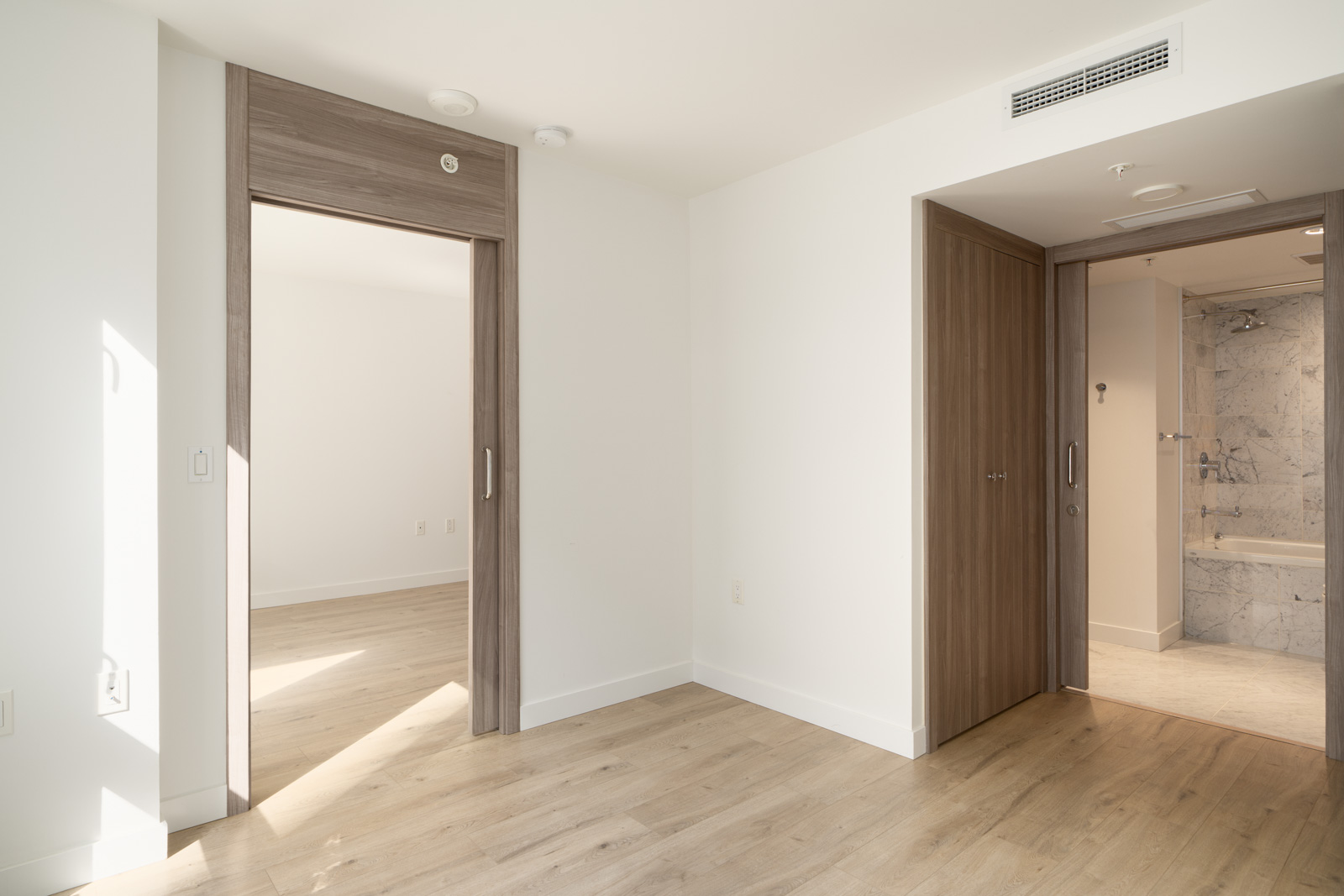 Empty modern room with light wood floors, white walls, two open wood doors, and a view into a tiled bathroom with bathtub in Vancouver.