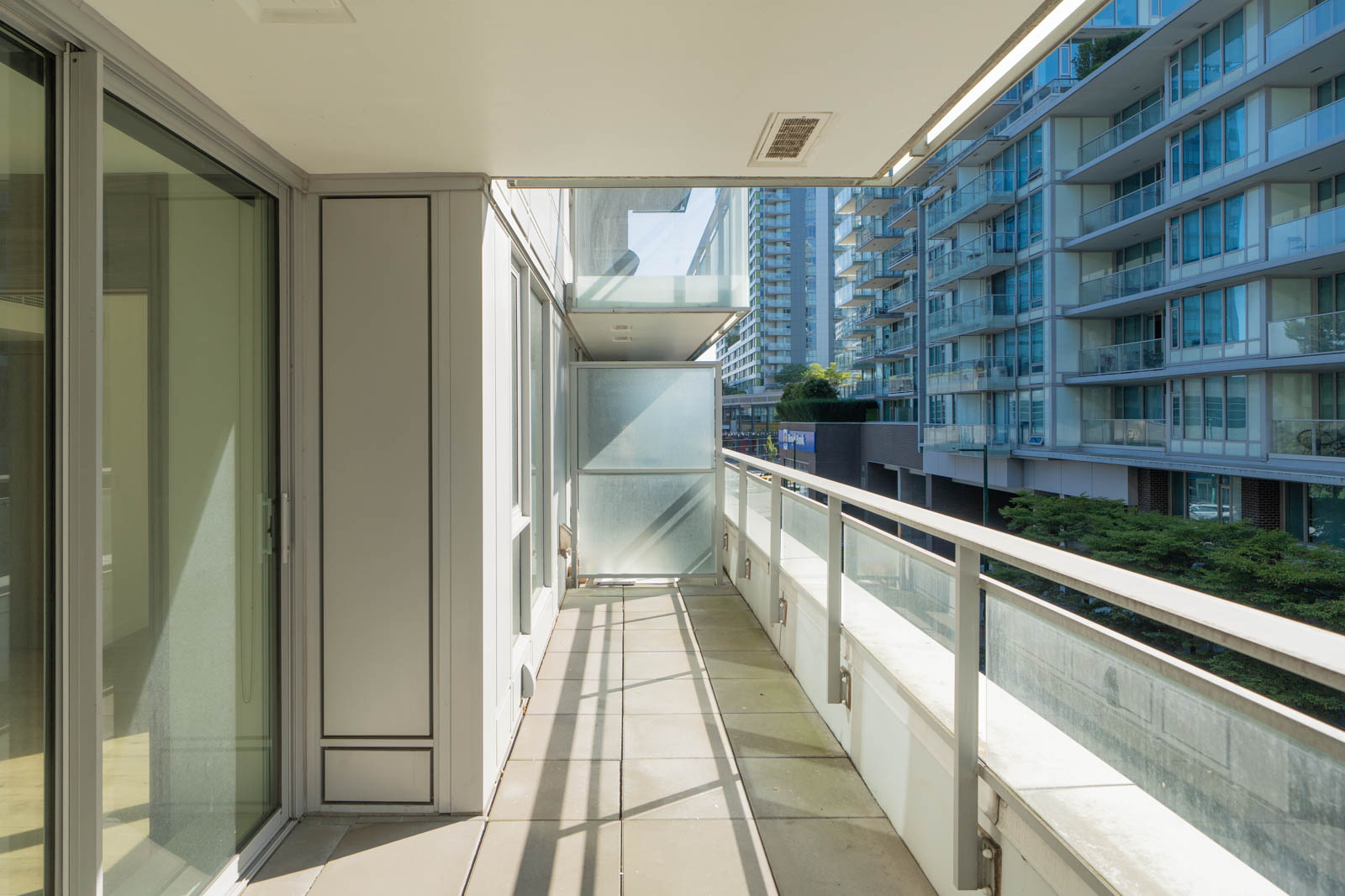Balcony with glass railing overlooks Marine Drive high-rises in Vancouver; sunlight casts shadows on tiled floor.