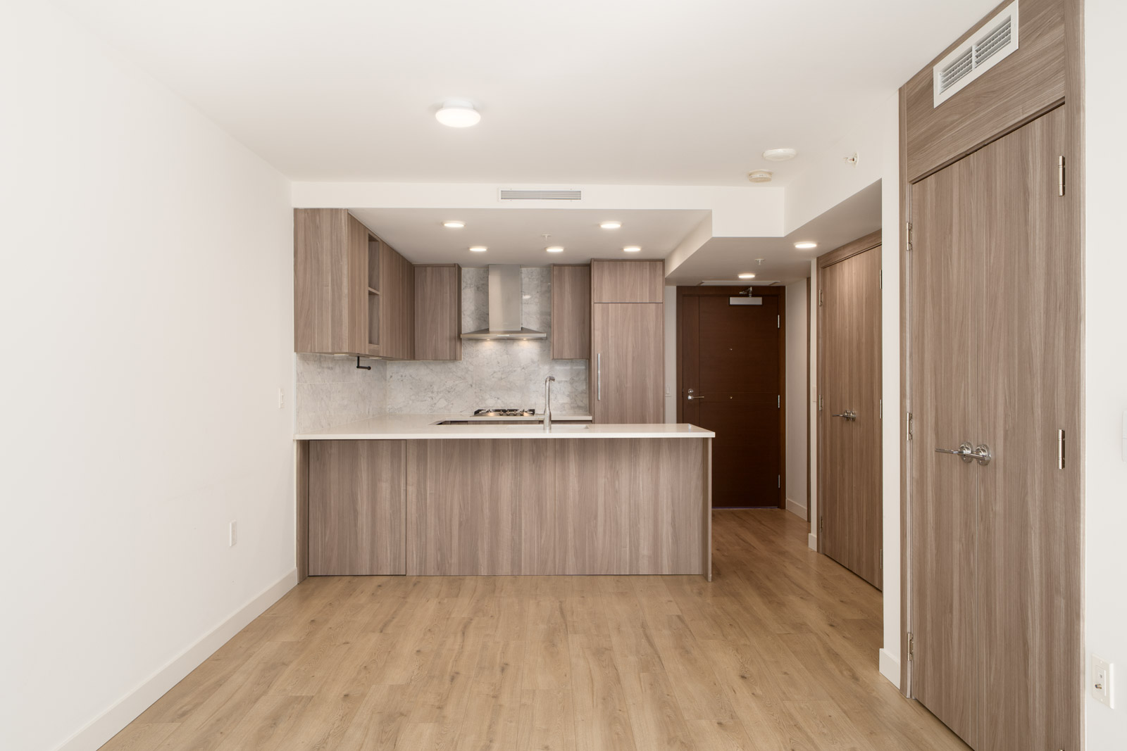 Modern Vancouver apartment kitchen with wood cabinets, marble backsplash, light wood floors; entry door and closet in background.