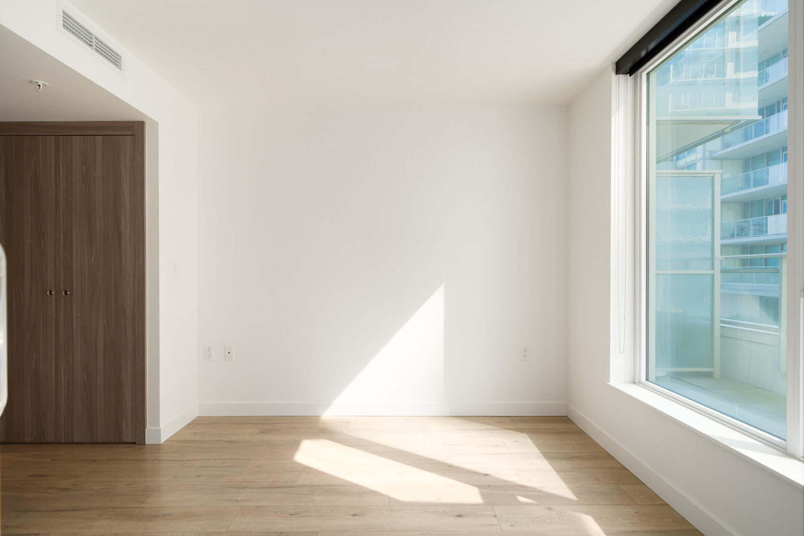 Sunlit empty room with light wood floors, white walls, closed closet left, large window right overlooking SW Marine Dr, Vancouver.