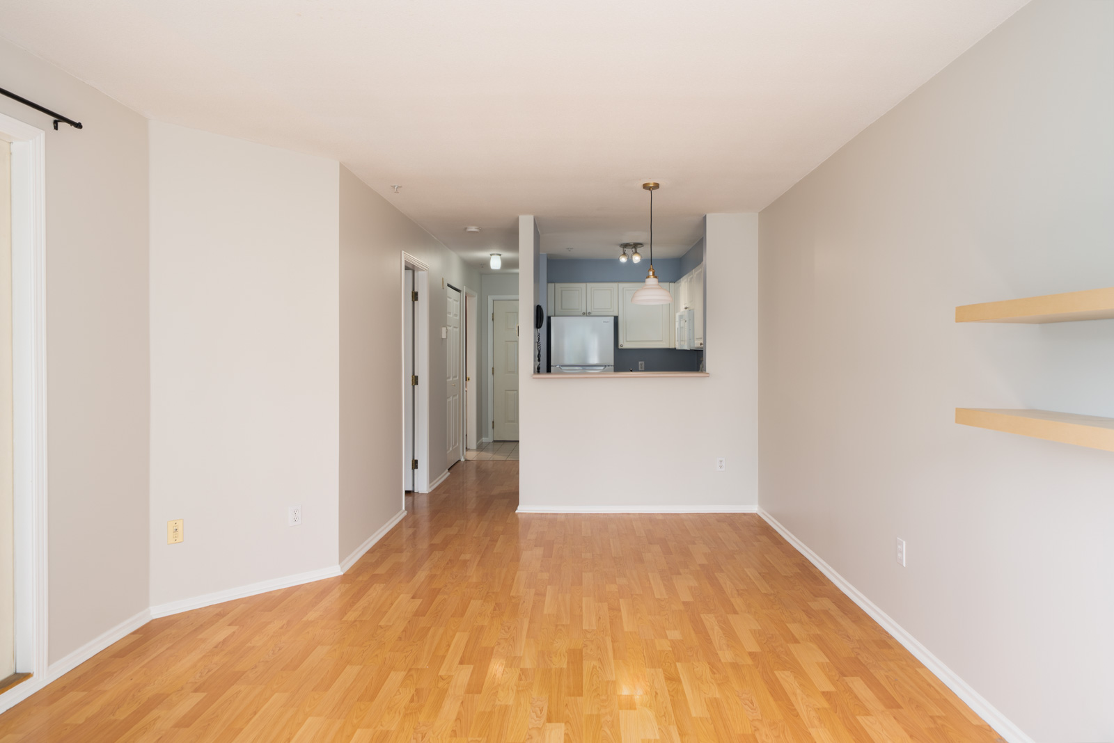 Empty living area with light wood floors, beige walls, built-in shelves, and view of small kitchen with white cabinets in Vancouver.