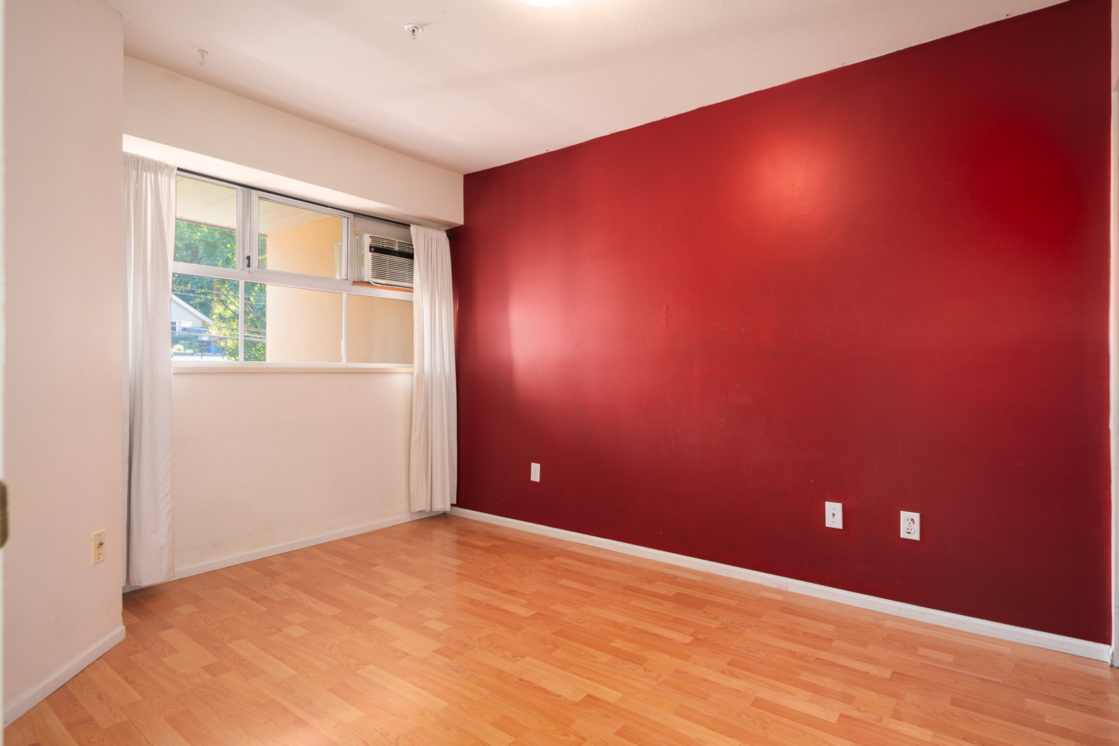 Bright empty room on Triumph St, Vancouver, featuring light wood floors, a red accent wall with outlets, and a window with white curtains.