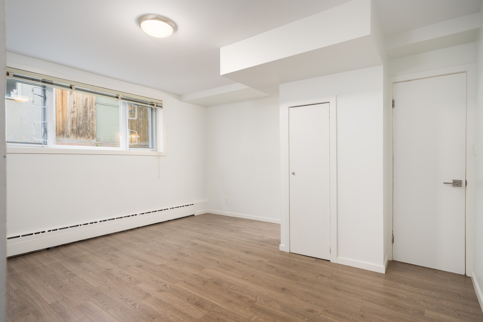 Empty room with light walls, large window with blinds, wood floor, closet and entry doors, ceiling light, and baseboard heater in Vancouver.