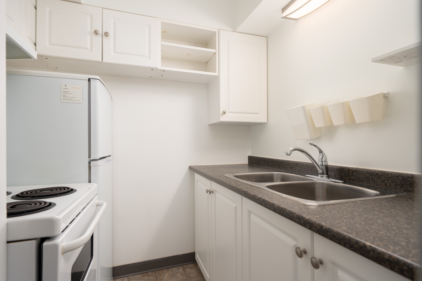 Compact lower-level Vancouver kitchen with white cabinets, dark counters, open upper doors revealing empty shelves, and bright lighting.