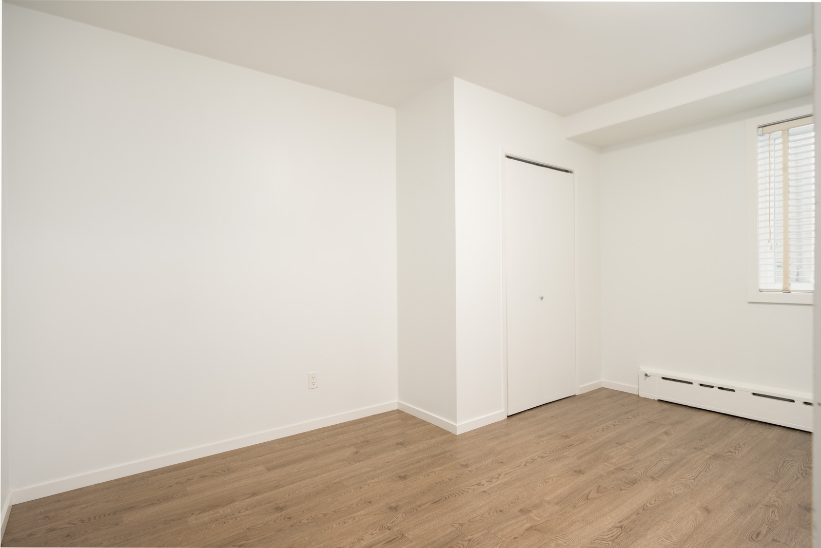 Empty room with white walls, wood floors, closed closet door, baseboard heater, and right-side window—Lower Floor, Vancouver.