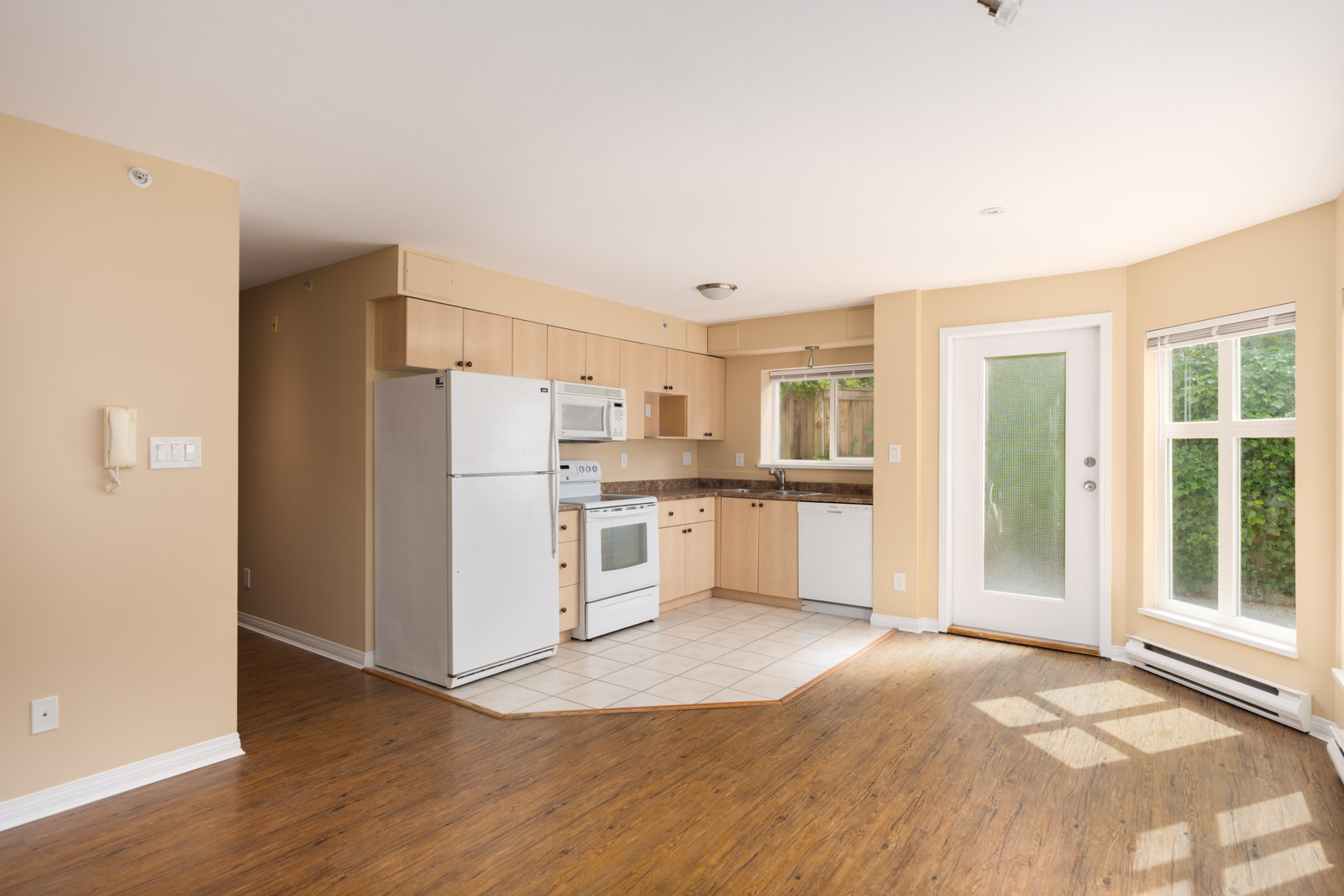 Small kitchen with white appliances and beige cabinets opens to a wood-floored living area with big windows and glass door in Vancouver.