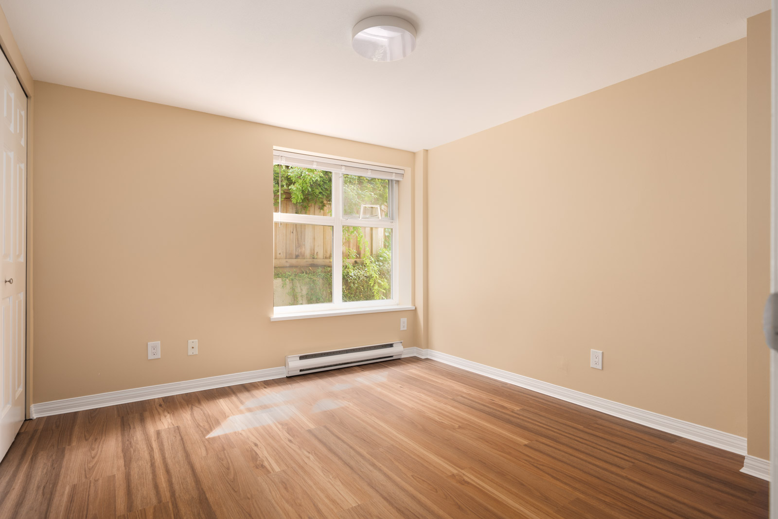 Empty room on 1st floor, 1099 West 71st Ave, Vancouver; beige walls, white trim, wood floor, sunny window, baseboard heater.