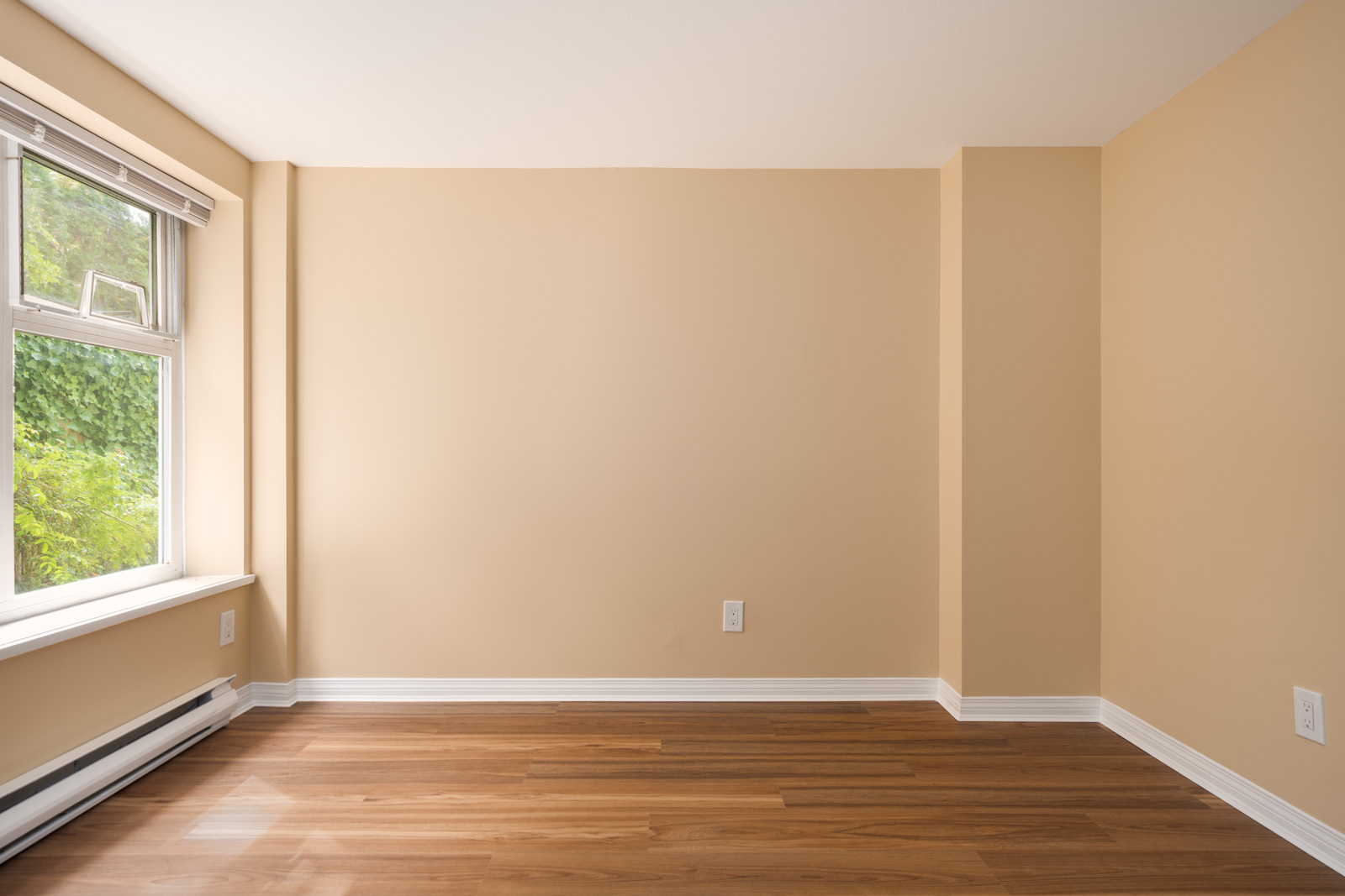 Empty beige room with light brown wood floors, white baseboards, and large left window letting in daylight at 1099 W 71st Ave, Vancouver.