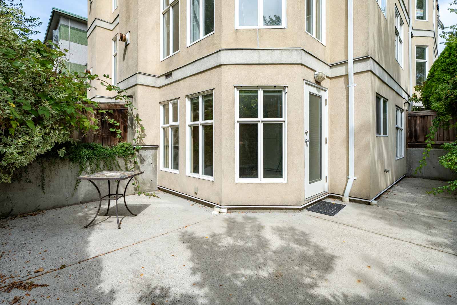 Concrete patio with a round table beside a beige building and glass door at 1099 West 71st Ave, Vancouver; shrubs and fence border edge.