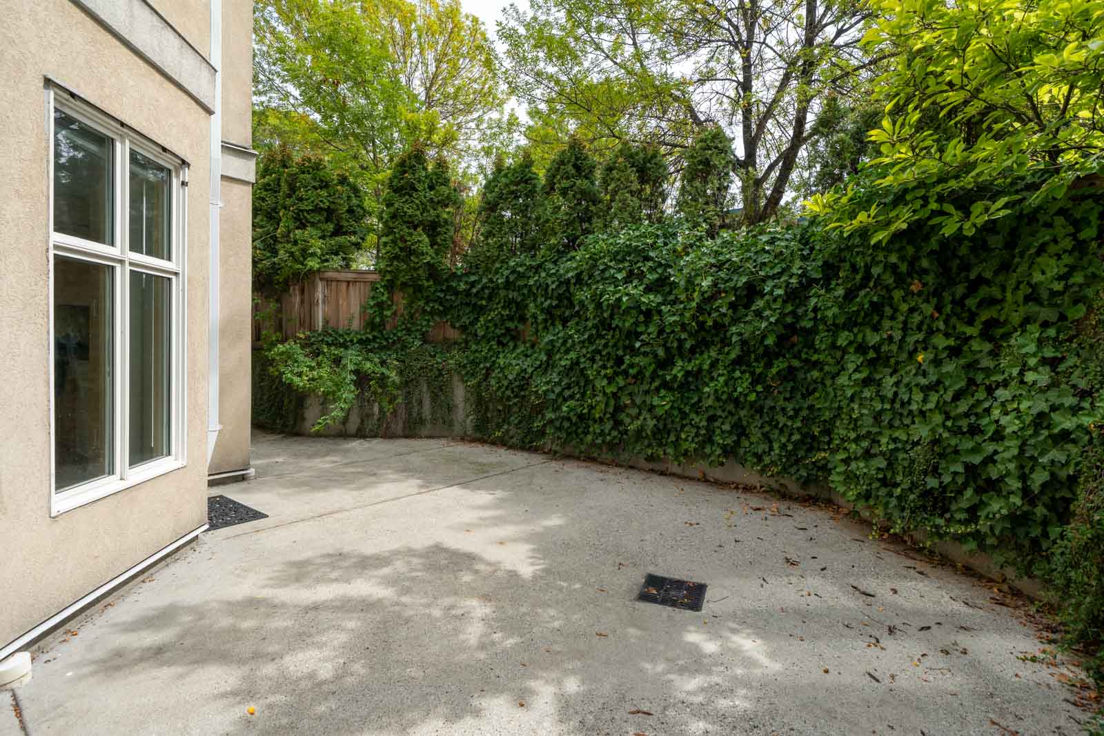 Concrete patio by a beige building at 1099 West 71st Ave, Vancouver, bordered by tall greenery and vine-covered fences.