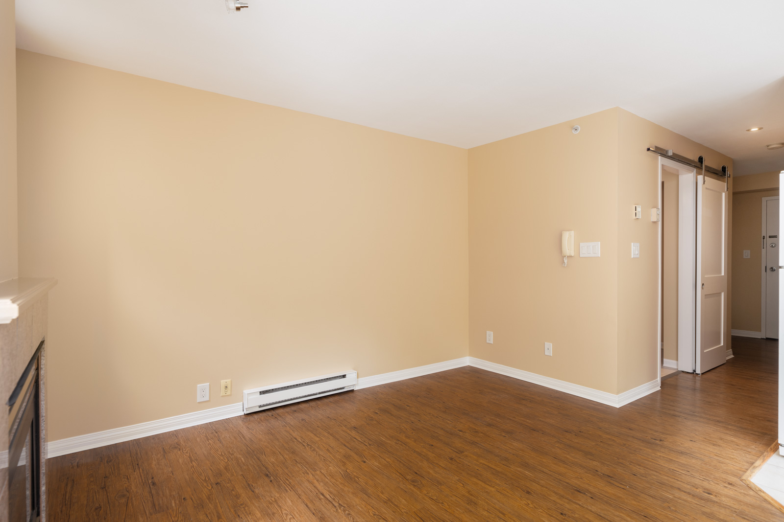 Empty room with beige walls, wood floor, baseboard heater, outlets, and a sliding barn door to a hallway in Vancouver.