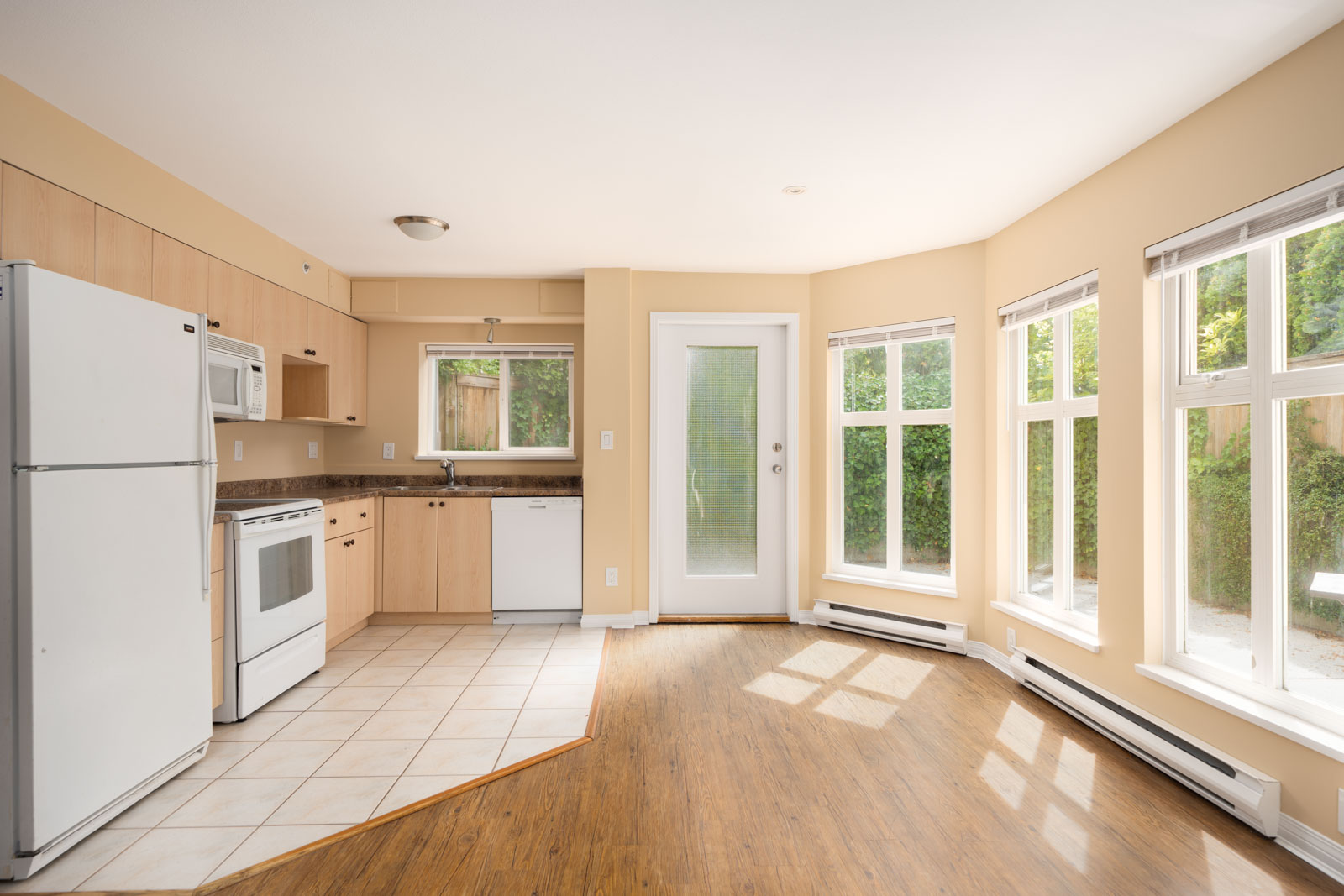 Bright kitchen with light wood cabinets, white appliances, tile and wood floors, large windows, and glass door to greenery; 1st floor, Vancouver.
