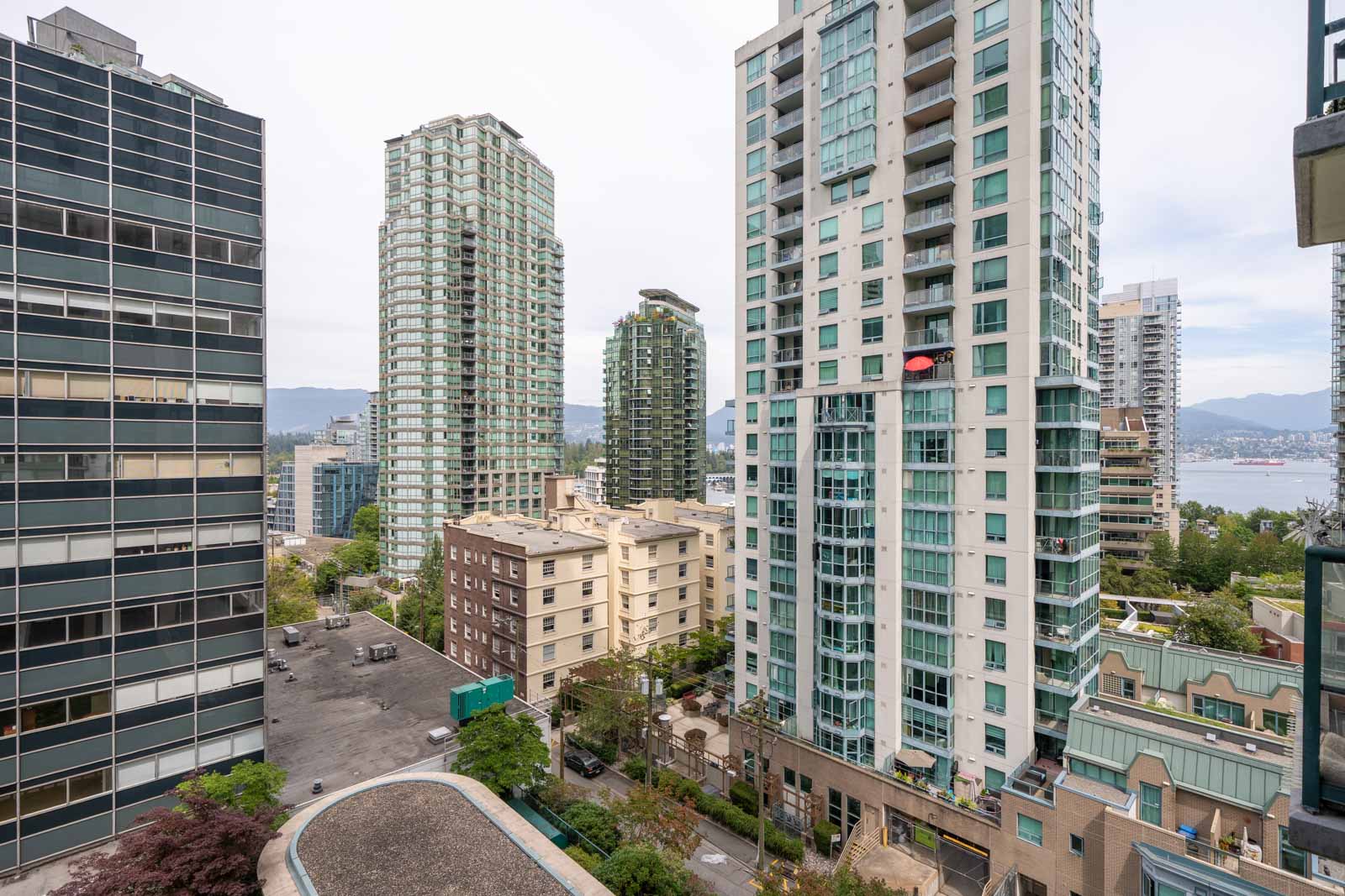 Modern high-rise apartments near 1239 W Georgia St, Vancouver, with smaller homes, distant water, and mountains under cloudy skies.