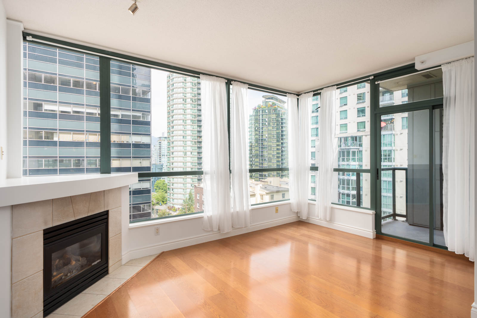 Corner room with wood floors, large windows, fireplace, sheer curtains, and glass door to a 7th-floor balcony overlooking Vancouver.