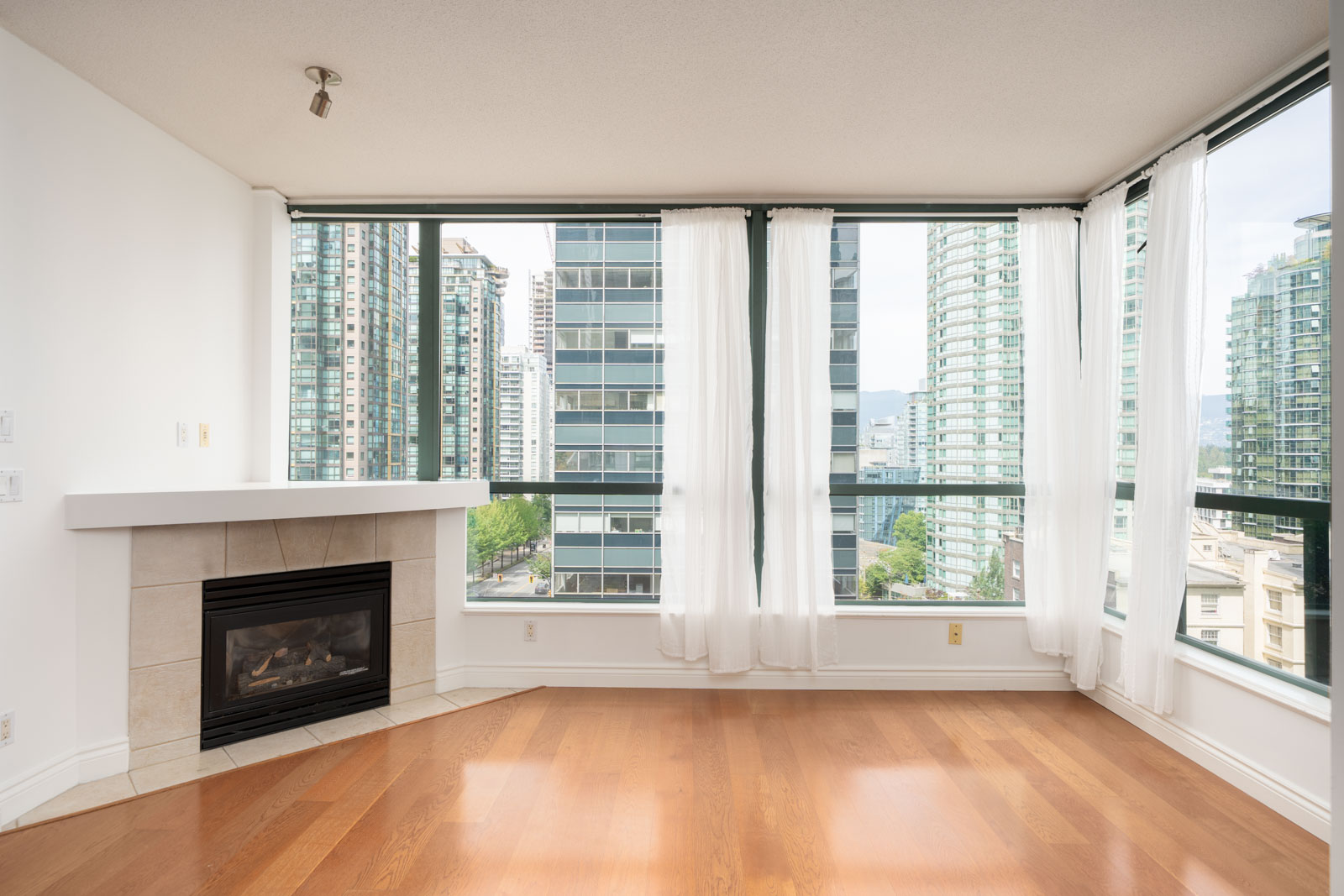 Corner room with hardwood floors, large windows, white curtains, modern fireplace, city view at 1239 W Georgia St, Vancouver.