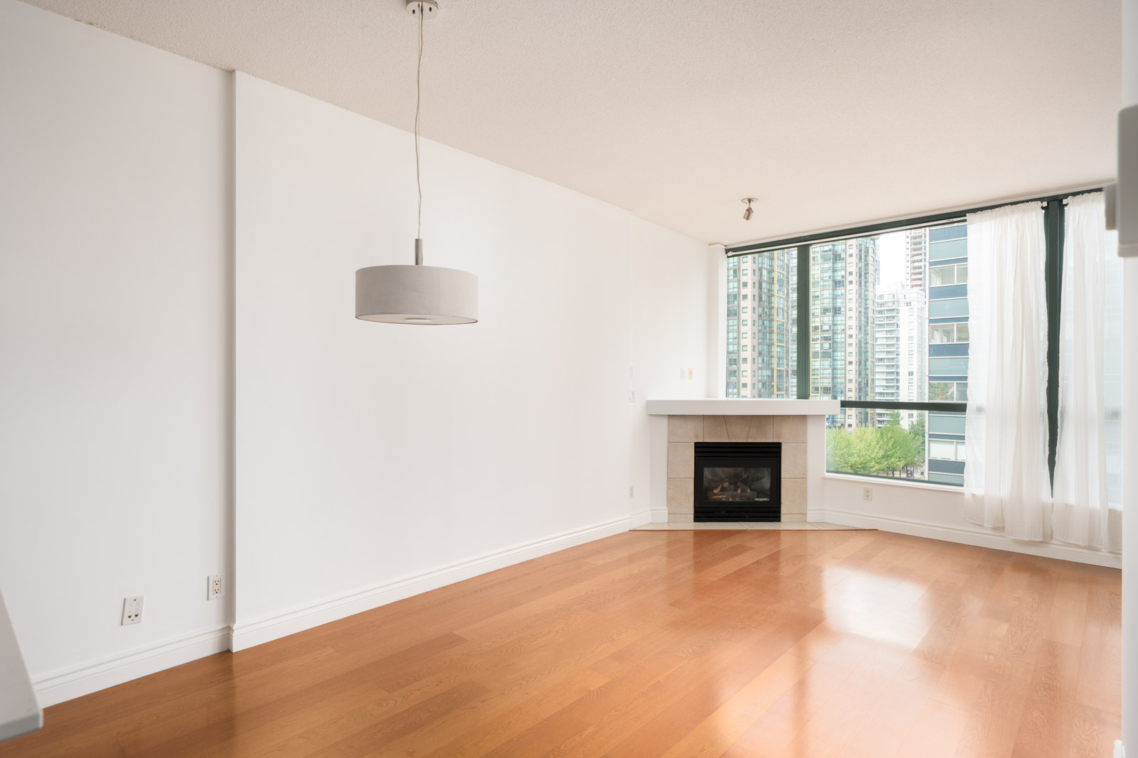 Modern living room with wood floors, white walls, ceiling light, fireplace, and large windows facing cityscape in Vancouver.