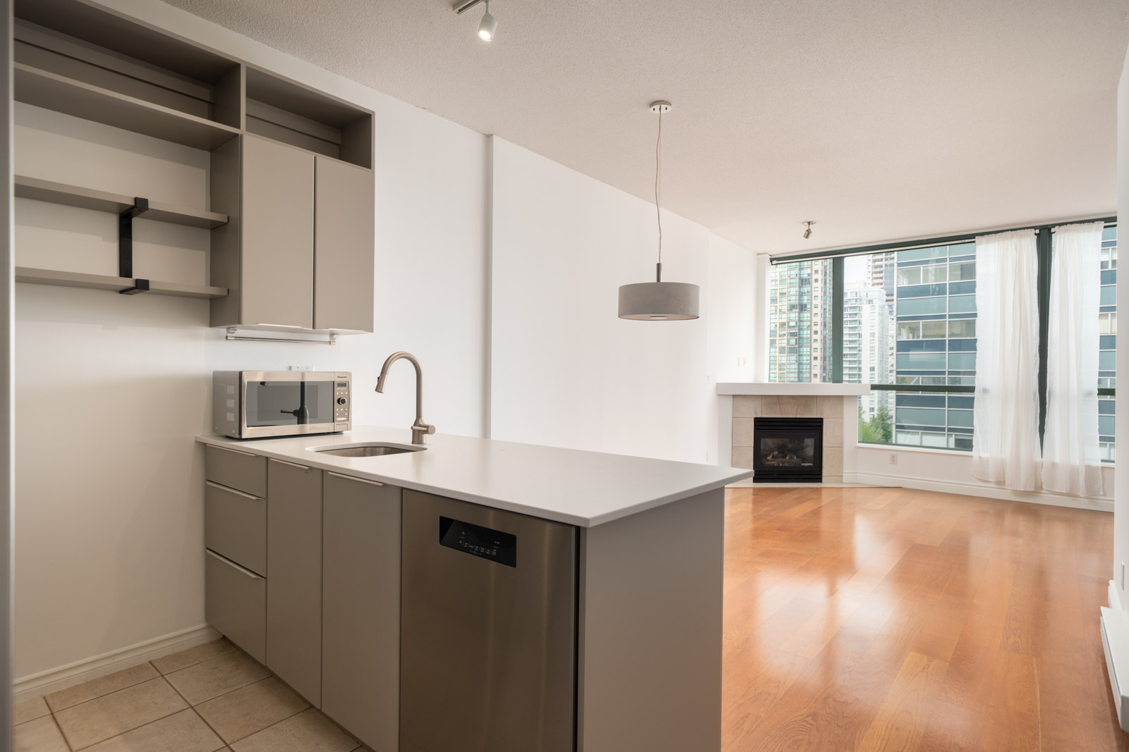 Modern apartment kitchen with gray cabinets, built-in sink, microwave, and dishwasher, opening to living area with wood floors and fireplace at 1239 W Georgia St, Vancouver.