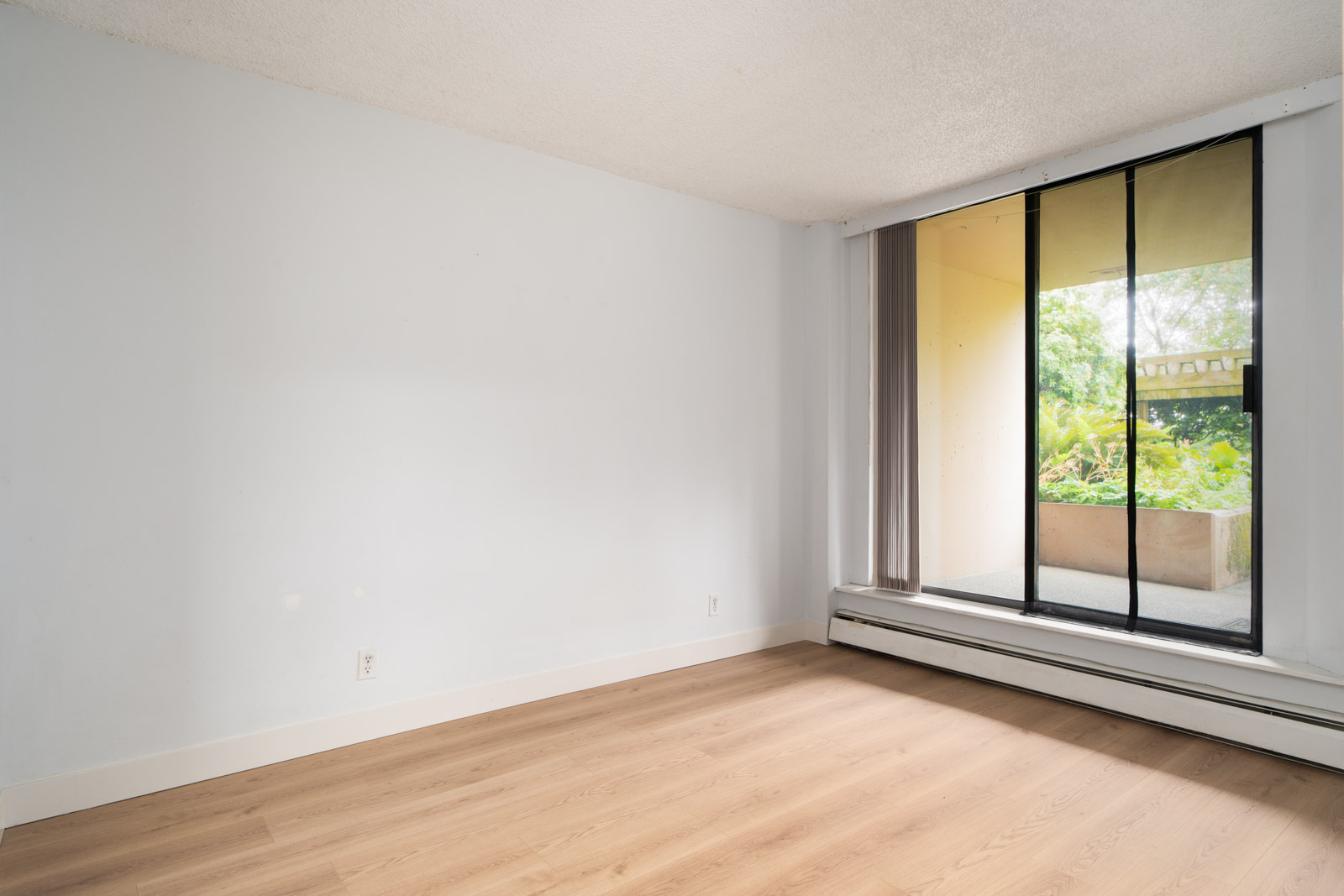 Empty room with light wood floors, white walls, and a sliding glass door to a green patio in a Coquitlam condo at 460 Westview St.