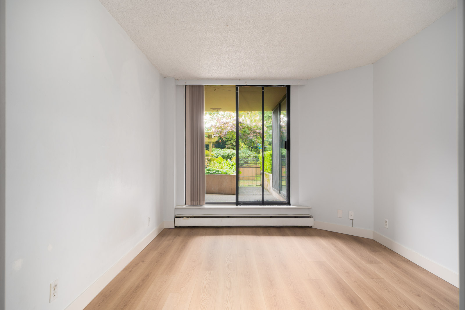 Empty room with light walls and wood floors, large glass sliding door to patio and garden. Located in a 1st floor condo in Coquitlam.