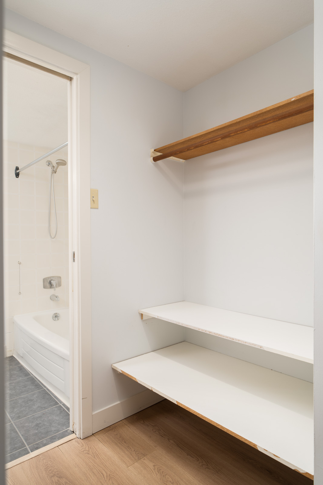 Empty closet with two white shelves and a wooden rod in a 1st floor Coquitlam condo, beside a bathroom seen through an open door.