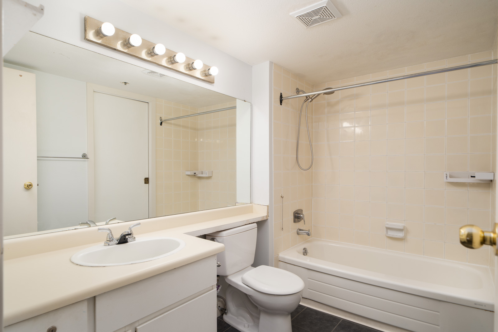 White bathroom in a 1st floor Coquitlam condo with a large mirror, single sink vanity, toilet, tub with shower, and beige tiled walls.