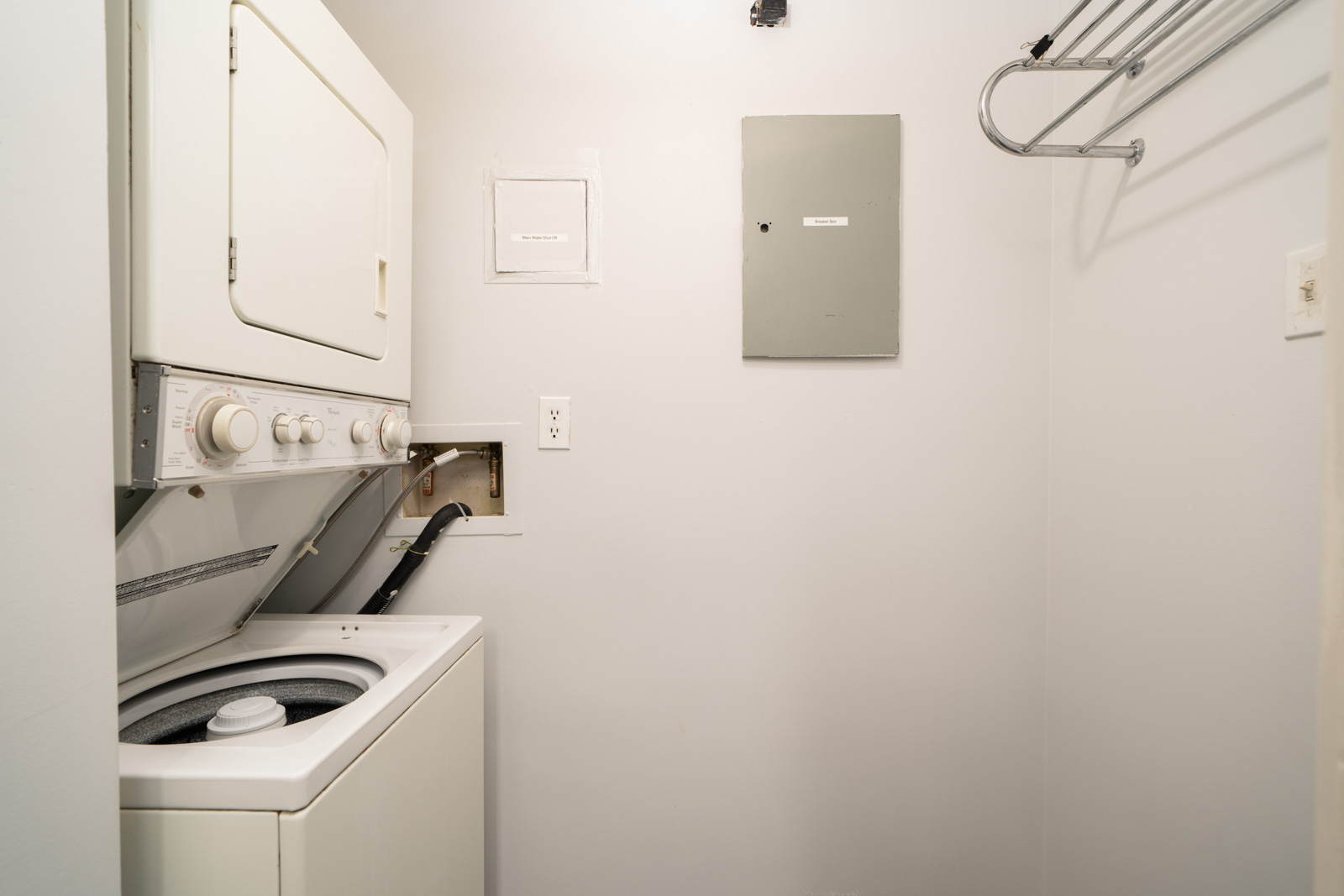 Compact laundry room in a 1st floor Westview St, Coquitlam condo with stacked washer/dryer, open washer lid, hoses, panel, and drying rack.