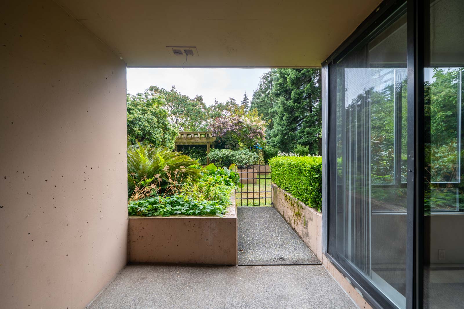 View from a covered patio at a 1st floor Westview St Coquitlam condo, overlooking a small garden with hedges and a metal gate.