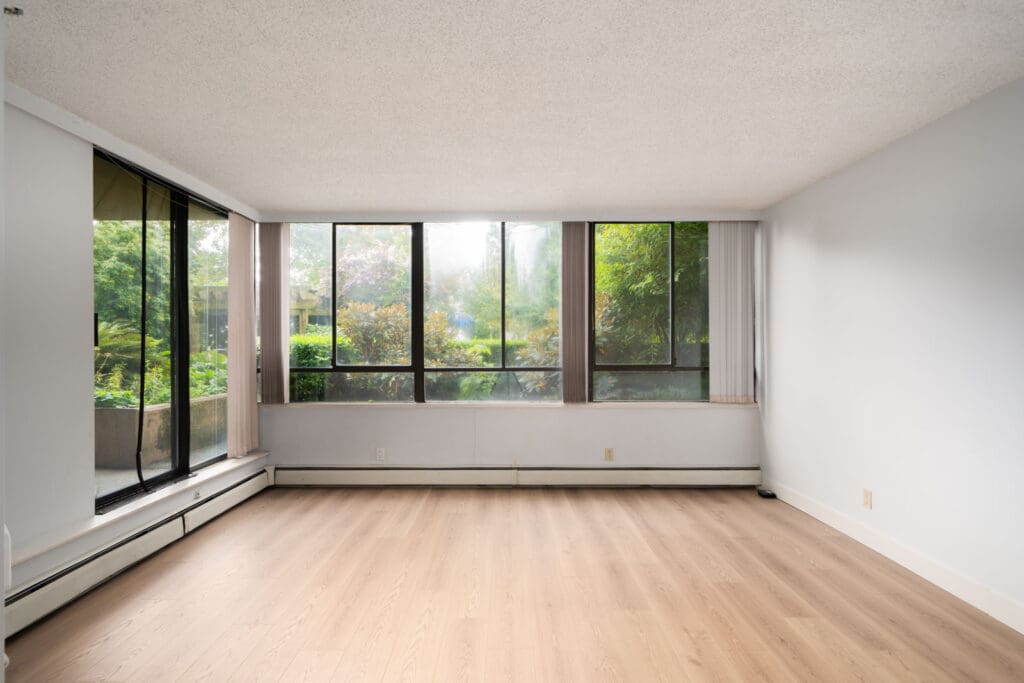 Empty room with light wood floor, white walls, large windows, and views of greenery in a Coquitlam 1st floor condo.