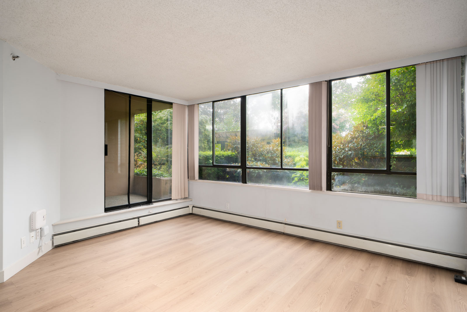 Bright, empty room with light wood floors, large windows, vertical blinds, and sliding glass door to greenery. Coquitlam condo.