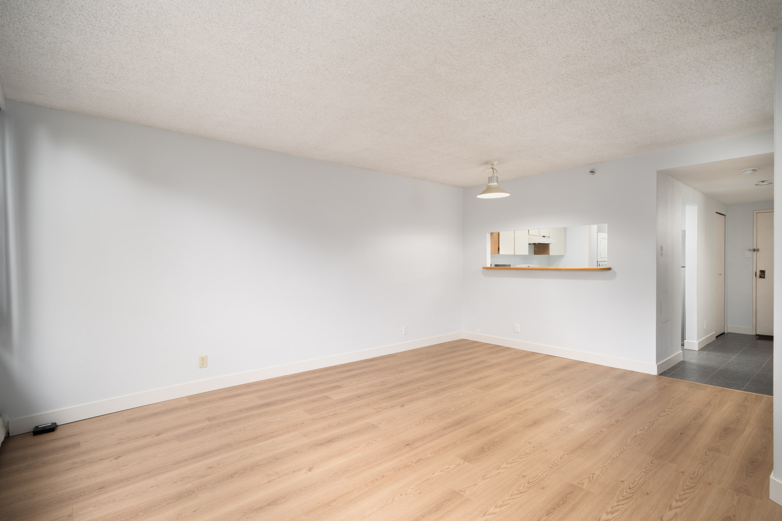Empty room with light wood floors, white walls, ceiling light, pass-through kitchen window, and entryway in a Coquitlam condo.