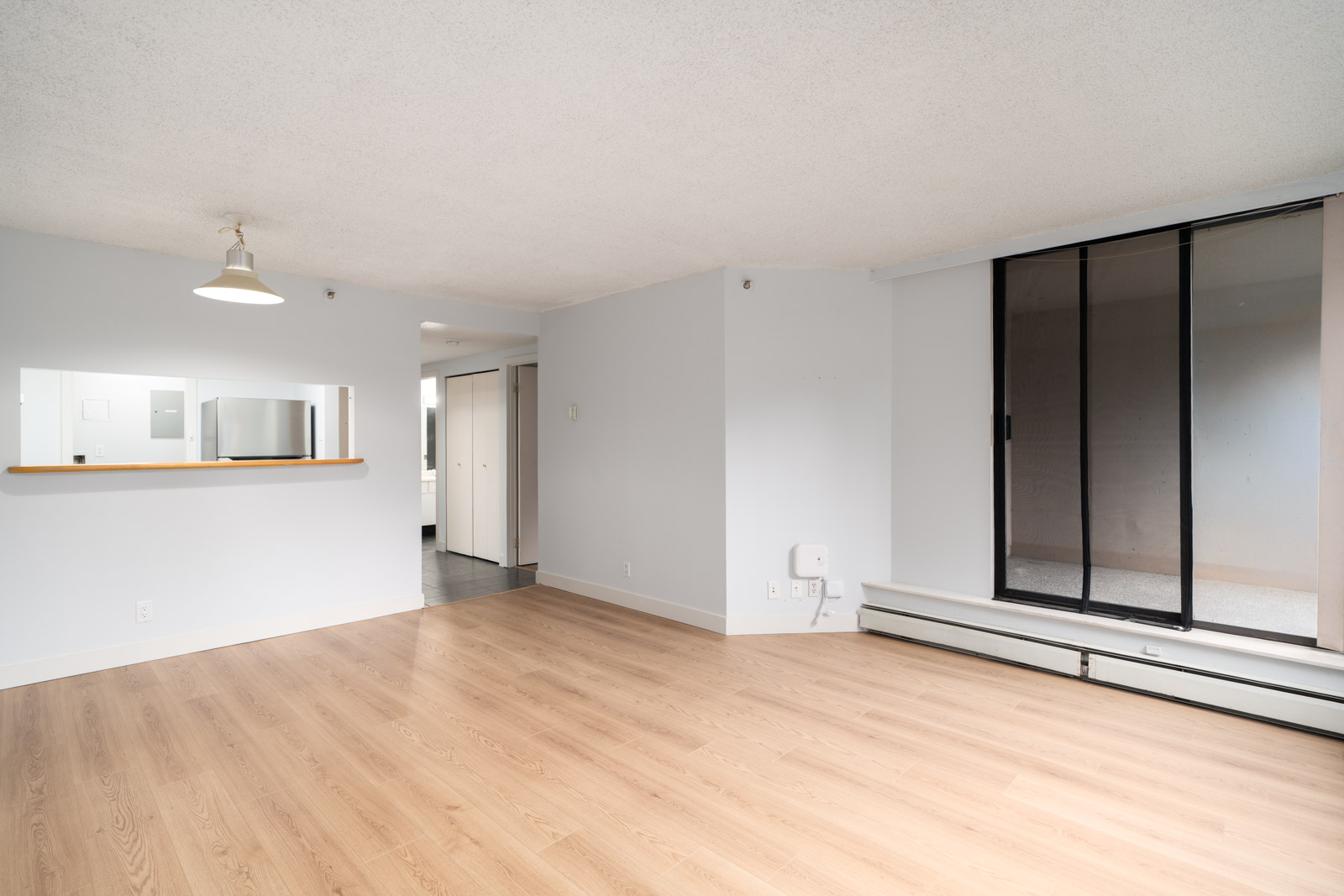 Empty living room with light wood floors, white walls, passthrough to kitchen, sliding doors, and hallway in Coquitlam condo.