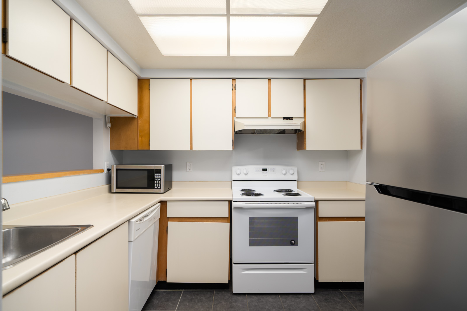 Compact kitchen in a 1st floor Coquitlam condo with white cabinets, stainless fridge, electric stove, and bright ceiling lights.
