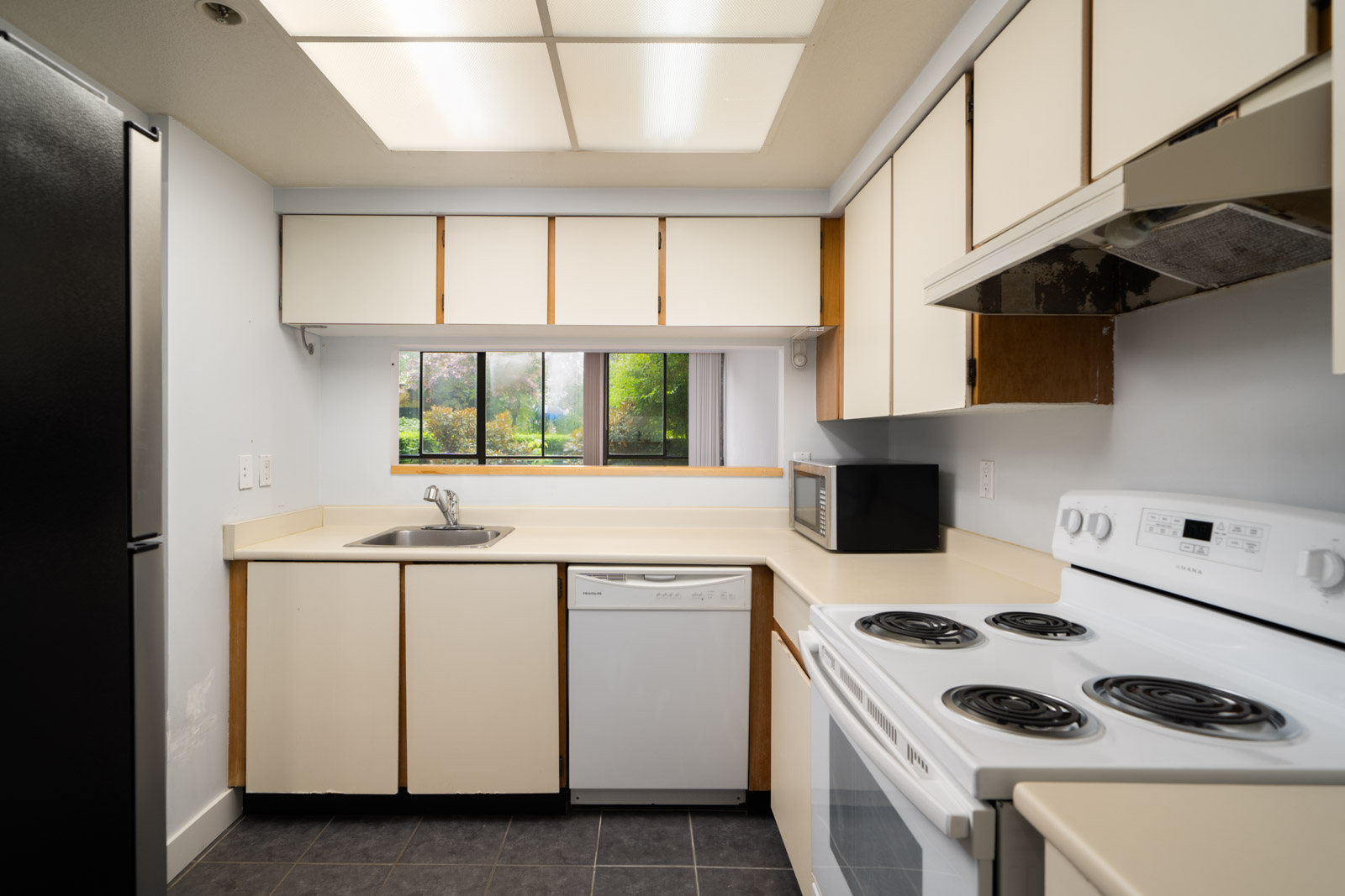 Small kitchen with white cabinets, electric stove, dishwasher, microwave, and a sink under a window with a green outdoor view in Coquitlam.