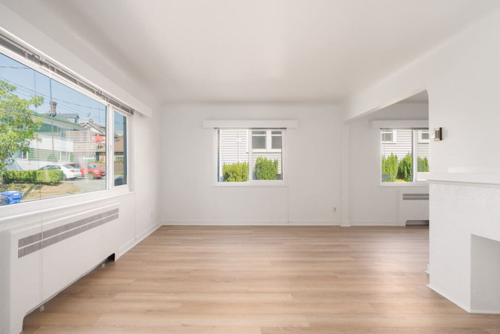Bright living room with wood floors, white walls, large windows, baseboard heater; overlooks cars and greenery on Royal Ave, New Westminster.