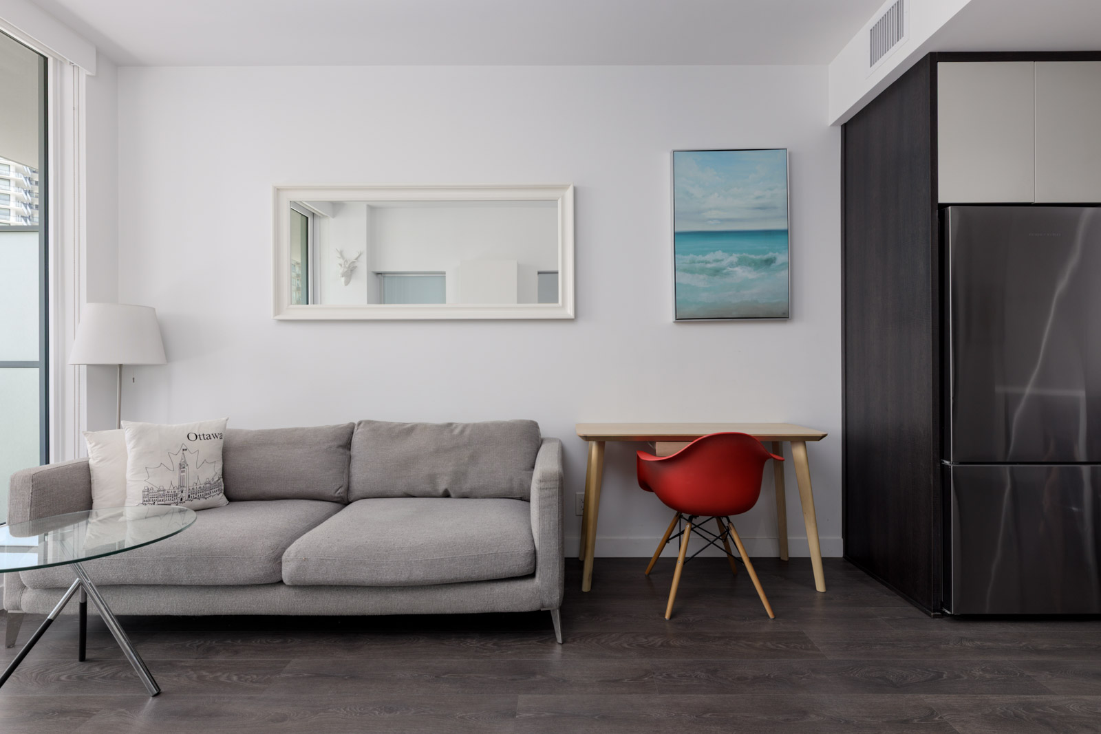 Modern living room at 567 Clarke Rd, Coquitlam with gray sofa, glass coffee table, red desk chair, wall art, and stainless fridge.