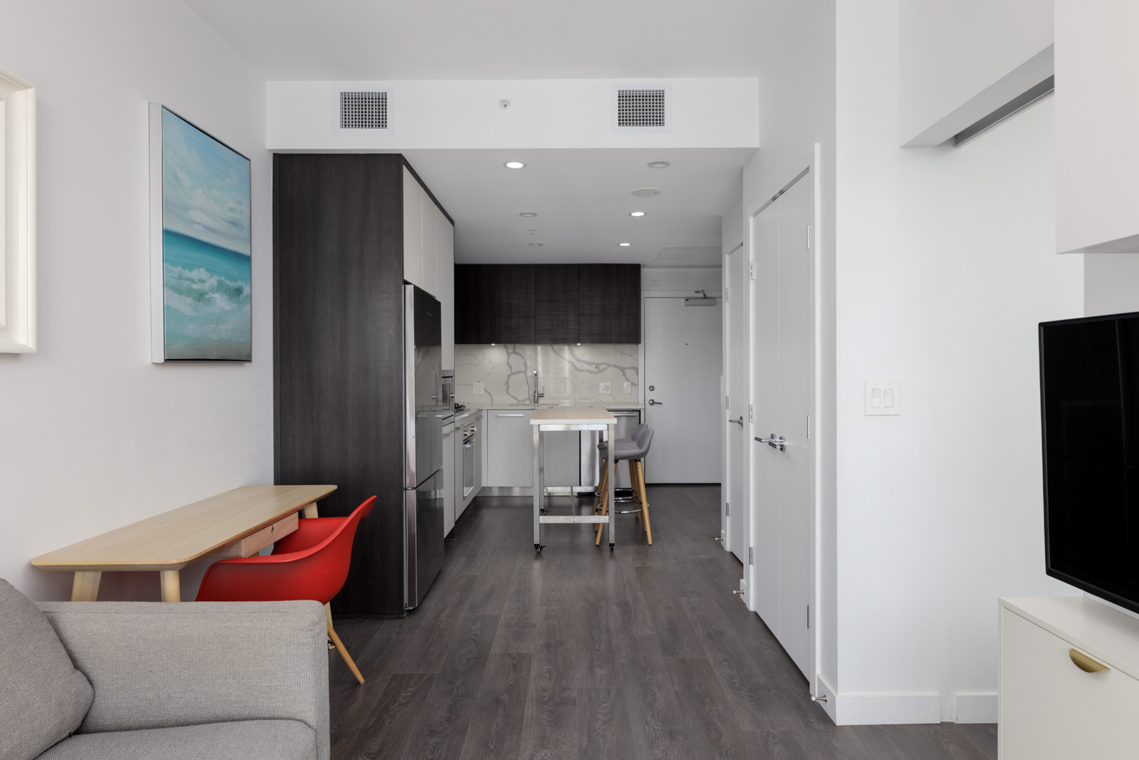 Modern apartment at 567 Clarke Rd, Coquitlam with dark wood floors, kitchenette, red chair, gray sofa, and ocean artwork.