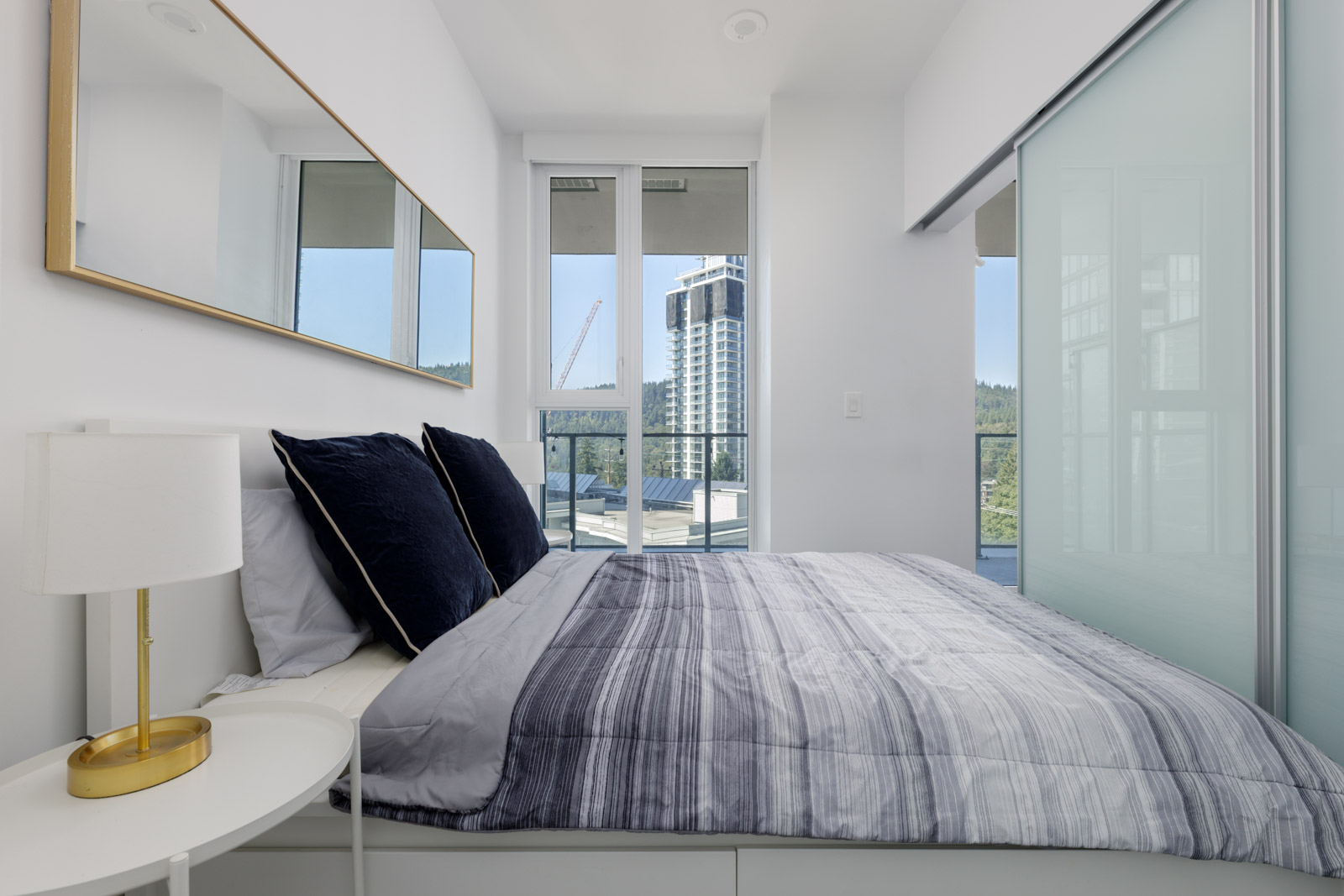 Modern bedroom with blue and gray bedding, white lamp, large mirror, and window overlooking 567 Clarke Rd in Coquitlam cityscape.