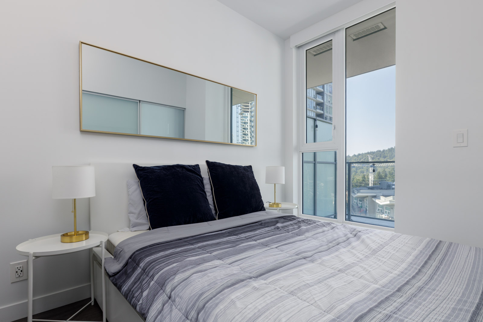 Modern bedroom in Coquitlam featuring a made bed, bedside tables with lamps, large mirror, and city views through tall windows.