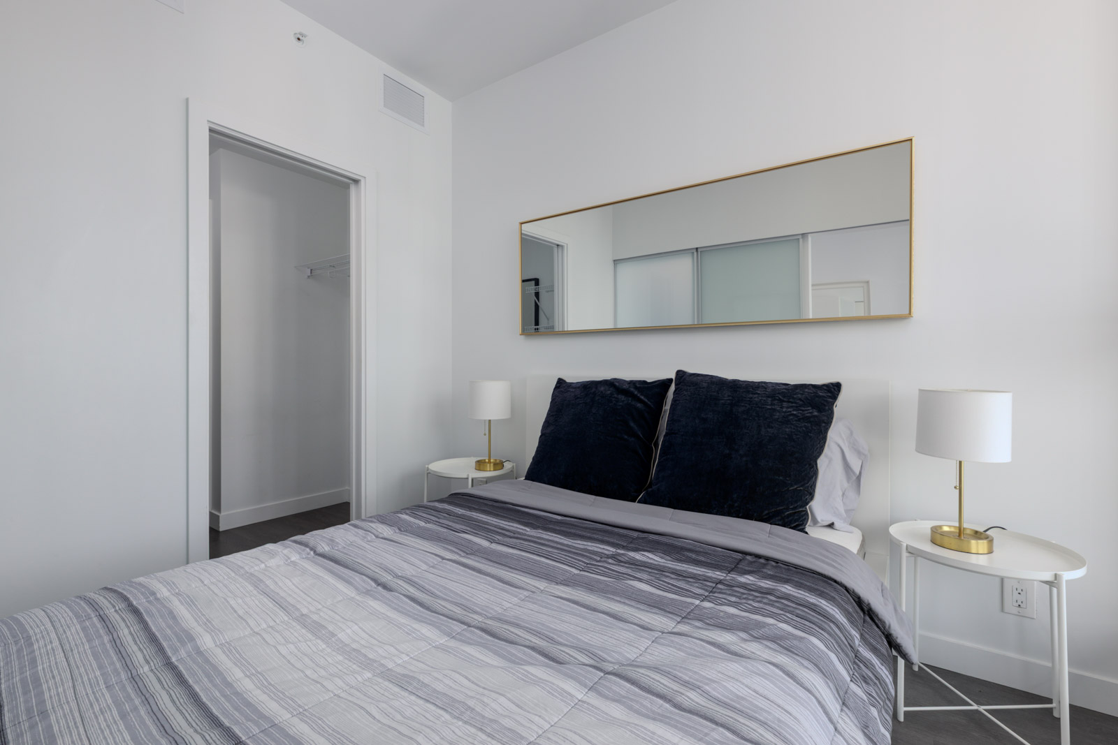 Minimalist bedroom at 567 Clarke Rd, Coquitlam with a tidy bed, black pillows, white side tables, lamps, and large mirror above. Doorway and closet in back.