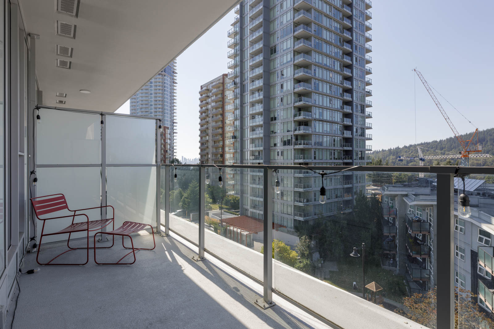 A modern balcony at 567 Clarke Rd in Coquitlam with two red chairs and glass railing, overlooking apartments and a construction crane.