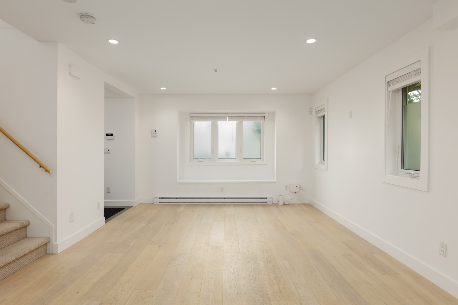 Empty room in Vancouver with light wood floors, white walls, three windows, recessed lighting, and a carpeted staircase on the left.