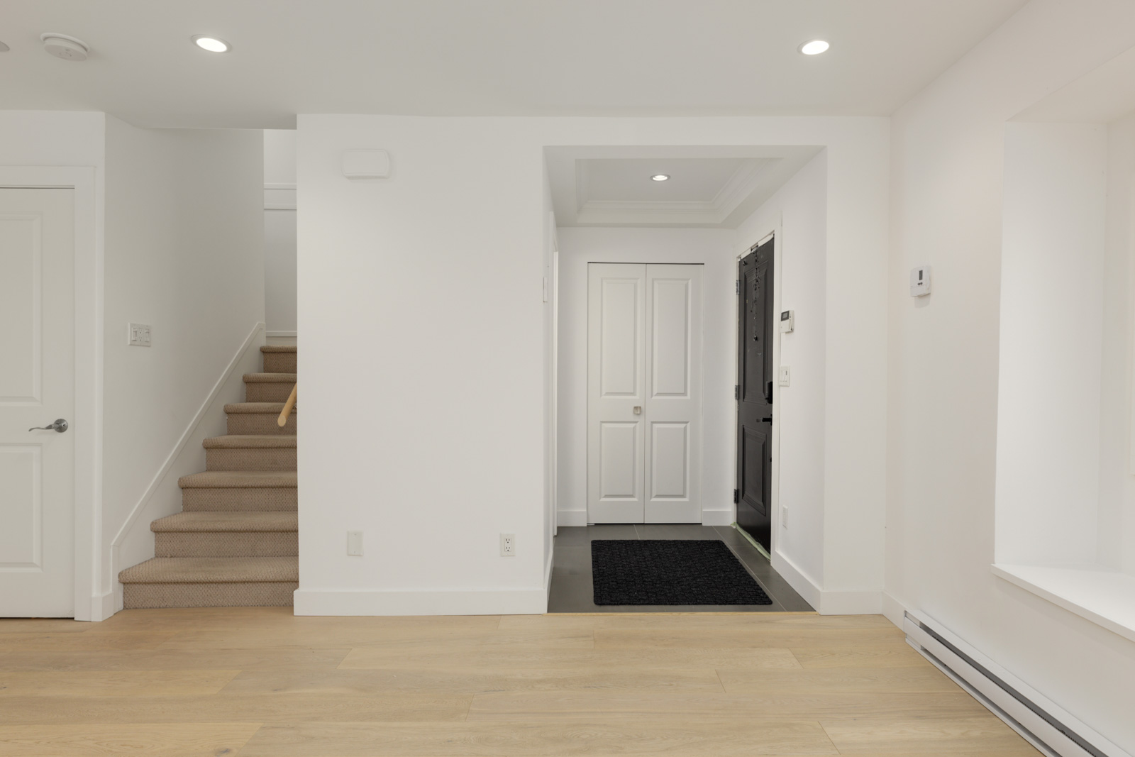 Modern entryway with light wood floors, white walls, black front door, double closets, and carpeted stairs in a Vancouver home.