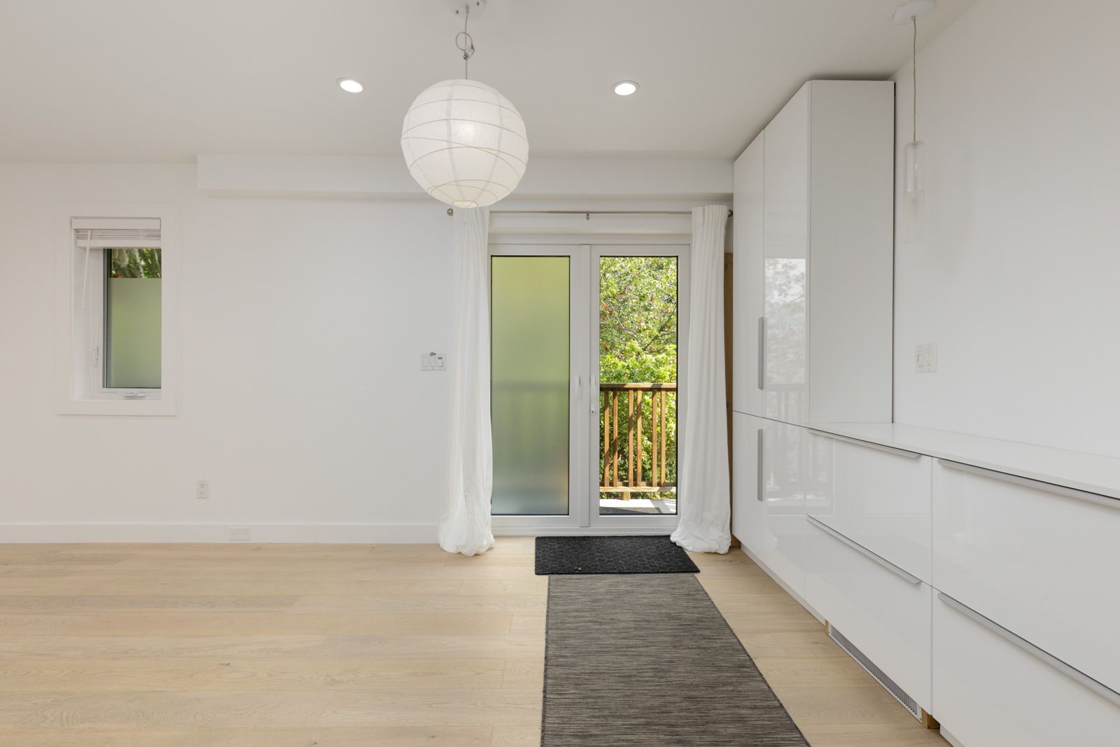 Minimalist room with light wood floor, white walls, round ceiling light, frosted glass door, white cabinets, and two dark rugs.